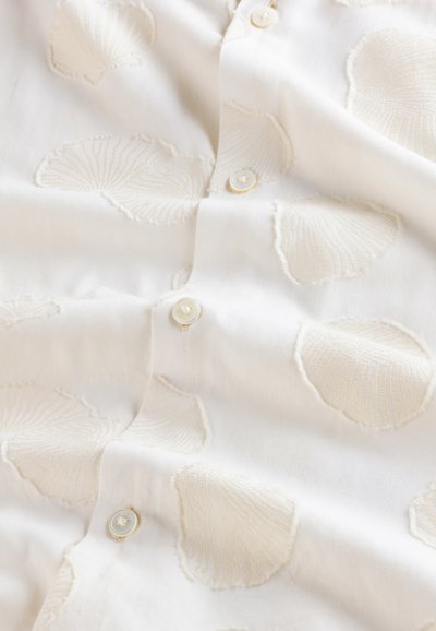Witte blouse met geborduurde schelp patronen. Voorzien van kleine, ronde knopen met een parelachtige afwerking. Zachte, gestructureerde stof met een subtiel ontwerp.