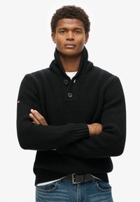 Schwarzer Strickpullover mit einem Schalkragen, ausgestattet mit gerippter Textur und zwei großen Knöpfen. Getragen über einem weißen Hemd, kombiniert mit blauen Jeans.