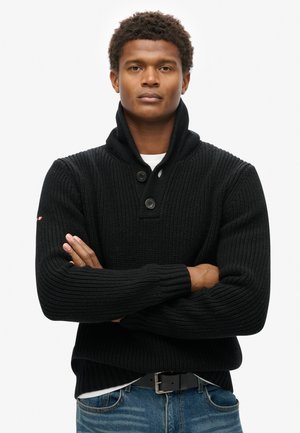 Zwarte gebreide trui met een sjaalkraag, voorzien van een geribde textuur en twee grote knopen. Draag het over een wit shirt, gecombineerd met blauwe jeans.