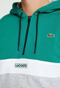 Sudadera con capucha de tela verde azulado, cierre de cremallera, cordones negros y una sección inferior blanca con el logo "LACOSTE" en verde.