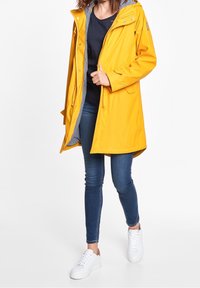 Gul vattentät regnjacka med huva, framzip och fickor; fodrad med randig tyg. Parat med blå jeans och vita sneakers.