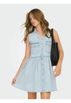 Fiatal nő vékony világoskék gombos farmerruhában, fekete válltáskát tartva, csukott szemmel és lebegő hajjal pózol.