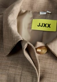 Bēša žaketes apkakle un atloks ar vienu brūnu pogu un dzeltenu JJXX zīmola etiķeti iekšpusē kakla izgriezumā.