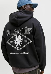 Sudadera negra con un gran gráfico de león blanco y el texto "GLACIER EST. 1990" en forma de diamante. La tela parece suave y tiene un corte relajado.