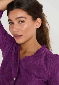 Blouse texturée violette avec un col en scallop, des manches bouffantes et un détail de bouton, fabriquée en tissu léger.