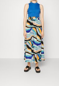 En blå, räfflad ärmlös topp som matchas med en flödande, flerfärgad mönstrad kjol med vågor av svart, blått, orange och aqua. Svarta sandaler.