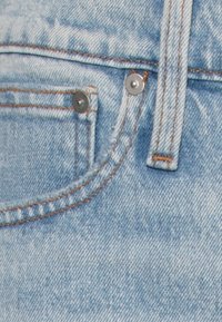 Jeans de denim azul claro em close-up, apresentando um pequeno bolso frontal com um botão prateado e costura laranja em contraste ao longo das costuras.
