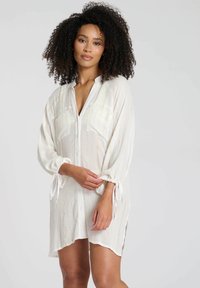 Camicia bianca leggera con un motivo testurizzato. Presenta maniche lunghe, legature regolabili ai polsini e due tasche frontali.