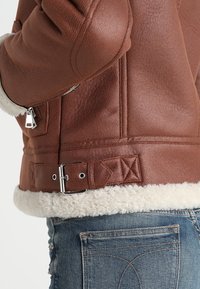 Giacca in finta pelle marrone con sottocollo in shearling, dotata di una fibbia in vita, tasca con zip e dettagli trapuntati vicino all'orlo.