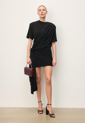 Donna in piedi contro un muro semplice, indossa un vestito nero asimmetrico e sandali neri con tacco alto, tiene una piccola borsa color bordeaux.