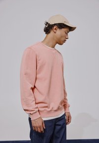 Jeune homme portant une casquette beige, un sweat-shirt rose et un jean bleu, debout contre un fond clair uni, la tête légèrement inclinée vers le bas sur le côté.