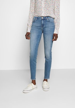 Person, die hellblaue Skinny-Jeans, ein weißes Langarmshirt mit einem mehrfarbigen Herzmuster und weiße Turnschuhe trägt, steht auf einem grauen Boden.