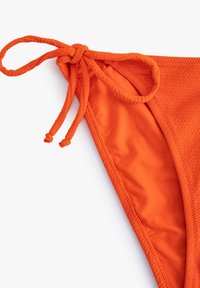 Parte inferior de bikini naranja con una tela texturizada y un forro suave, lazos ajustables con acentos trenzados y un diseño de corte triangular.