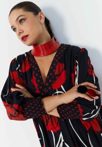 Bloemmotief jurk met zwarte basis, rode bloemen en stippen. Beschikt over een diepe V-hals, geplooide mouwen en een gestructureerde rode choker.