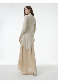 Beige lange mouwen top met asymmetrische zoom, gecombineerd met een vloeiende beige rok. Glad, lichtgewicht materiaal. Model kijkt van de camera weg.