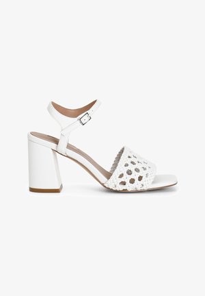 Las sandalias blancas de tacón cuentan con un tacón bloque grueso, diseño de tiras trenzadas y hebilla ajustable en el tobillo. Cuero suave y acentos texturizados.