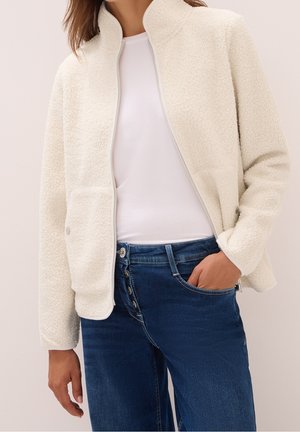 Femme portant une veste blanche texturée zippée sur une chemise blanche et un jean bleu à boutons, la main dans la poche.