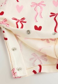 Bombažno oblačilo s kremasto ozadje, okrašeno z rdečimi in rožnatimi vzorci src in pentelj. Robovi so opremljeni z varnostnimi zaponkami.