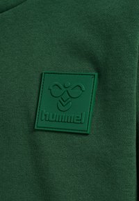 Tissu vert avec un patch en caoutchouc surélevé montrant un logo d'abeille stylisé et le texte « hummel » cousu sur la surface.