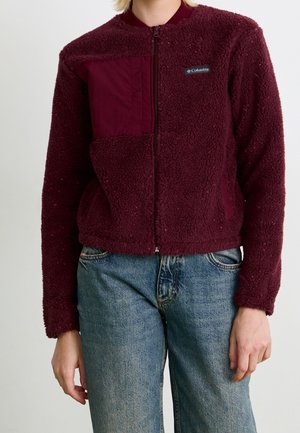 Personne portant une veste polaire zippée bordeaux avec une poche poitrine et un logo Columbia, associée à un jean bleu.