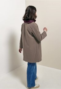 Abrigo de lana marrón claro con un corte holgado, que presenta mangas largas y bolsillos laterales, llevado sobre una blusa morada con cuello y combinado con unos vaqueros acampanados azules.