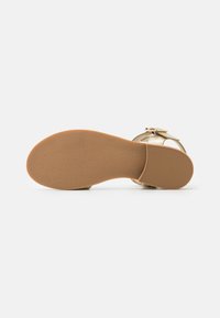 Sandal oro con una suola testurizzata, caratterizzata da un design piatto e una fibbia laterale. Il fondo ha un motivo di grip in gomma per una migliore aderenza.
