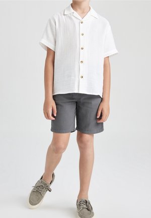Barn iført en hvid kortærmet skjorte med knapper, grå shorts og grå snøresko, der står på ét ben.
