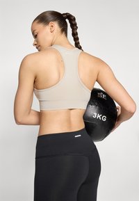 Bežs sporta krūšturis ar sacīkšu aizmuguri, apvienots ar melnajām augstajām legingi. Individuāls tur melnu, teksturētu 3kg medicīnas bumbu.