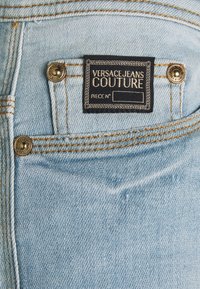 Vaqueros de mezclilla azul claro con una etiqueta que dice "VERSACE JEANS COUTURE". Presenta botones metálicos dorados y costuras naranjas a lo largo de las costuras.
