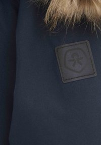 Marineblauw stof met een gestructureerd oppervlak, voorzien van een zwart rechthoekig label met een reliëflogo en een bontafwerking aan de bovenrand.