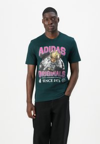 Green cotton t-shirt featuring large pink "ADIDAS ORIGINALS" graphic with a trophy and globe design on the front, crew neck, short sleeves.
