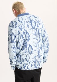 Långärmad skjorta i ljusblått med ett blått ormmönster, med krage och ribbade ärmslut. Tyget verkar mjukt och strukturerat.