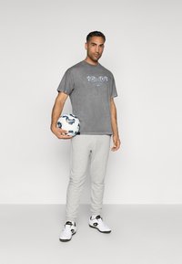 T-shirt cinza de algodão com estampado gráfico, calças de moletom cinzas e ténis brancos de desporto. A segurar uma bola de futebol com padrão. Vestuário atlético simples e casual.