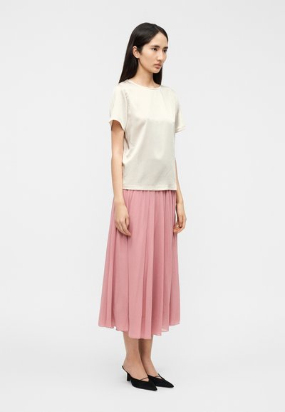 Μπεζ κοντομάνικη μπλούζα με στρογγυλή λαιμόκοψη, σε συνδυασμό με ροζ μακριά πλισέ φούστα και μαύρα mule με τακούνι. Ελαφρύ ύφασμα και λεία υφή.