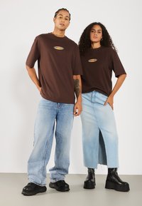T-shirts oversize en coton marron avec un logo, associés à un jean en denim bleu clair et une jupe, dotée d'une fente sur le côté, et des bottes à plateforme noires.