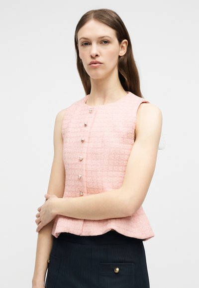 Γυναίκα που φοράει ροζ τοπ χωρίς μανίκια με υφή και κουμπιά από πέρλες, συνδυασμένο με σκουρόχρωμη φούστα με χρυσή λεπτομέρεια τσέπης κουμπιού.