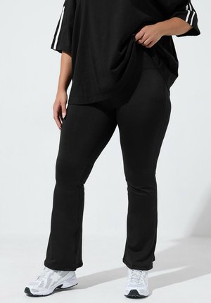 Person iført sorte vide leggings, sort oversize top med hvide striber på ærmerne og hvide sportssko, stående på en ensfarvet baggrund.