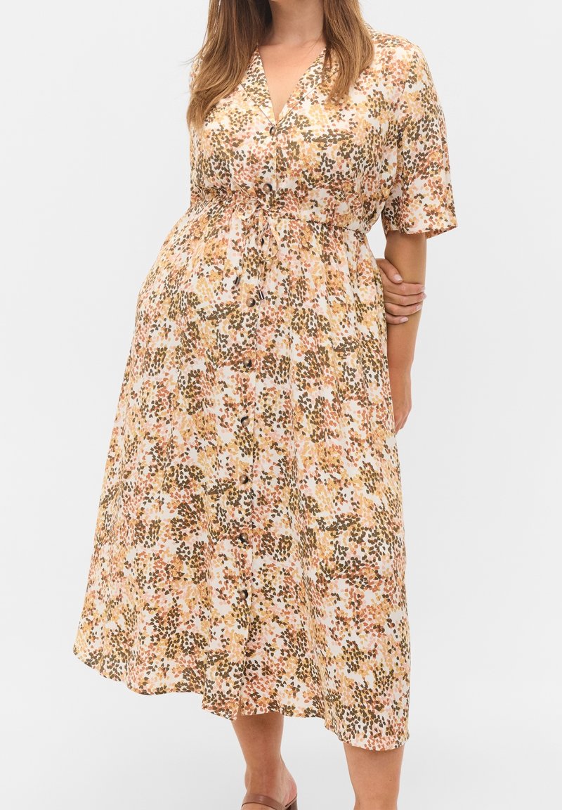 Femme portant une robe midi boutonnée à manches courtes avec un motif floral abstrait jaune, marron et rose et une taille cintrée.