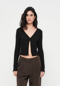 Μαύρο κοντό ζακέτα από υφασμάτινη υφή, με βαθύ V-λαιμόκοψη και κούμπωμα με ένα κουμπί, με κυματιστό τελείωμα και μακριά μανίκια.