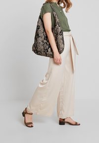 Gran bolsa de tote negra y beige con un patrón, textura suave, combinada con una blusa verde, pantalones de pierna ancha beige y sandalias marrones de tacón.