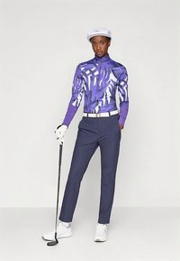 Γυναίκα σε μοτίβο μωβ μακρυμάνικο πουκάμισο γκολφ, μπλε παντελόνι, άσπρο γάντι, καπέλο και παπούτσια, κρατώντας ένα μπαστούνι γκολφ, στέκεται μπροστά σε απλό φόντο.