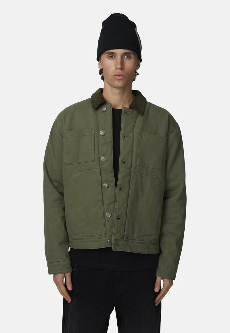 Veste en toile verte avec un col en velours côtelé, fermeture à boutons et deux poches poitrine. Portée sur une chemise noire et associée à un pantalon sombre.