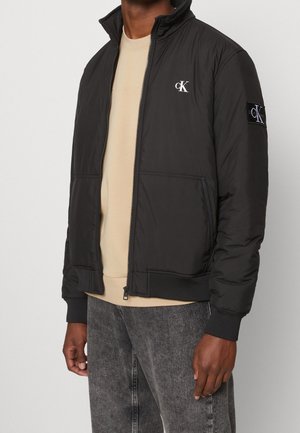 Homme portant une veste noire zippée avec le logo cK sur la poitrine et la manche, chemise beige en dessous et jean gris délavé.