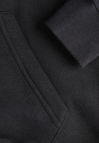 Sudadera negra con tejido que presenta un puño acanalado y un bolsillo tipo canguro. La textura es suave con un ligero estiramiento y las costuras están bien cosidas.