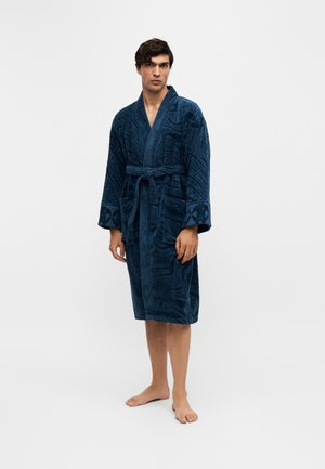 Jeune homme debout, pieds nus, portant un peignoir bleu foncé texturé, longueur genoux, avec une ceinture nouée à la taille, sur un fond clair uni.