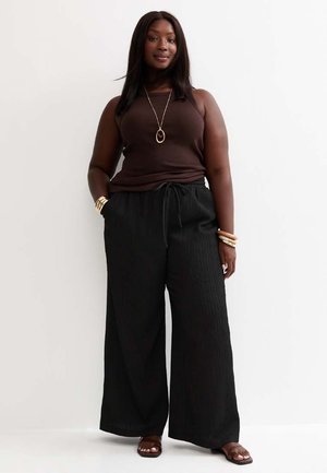 Vrouw staat tegen een witte muur, draagt een donkerbruine mouwloze top, zwarte wijde broek, sandalen, gouden ketting en armbanden.