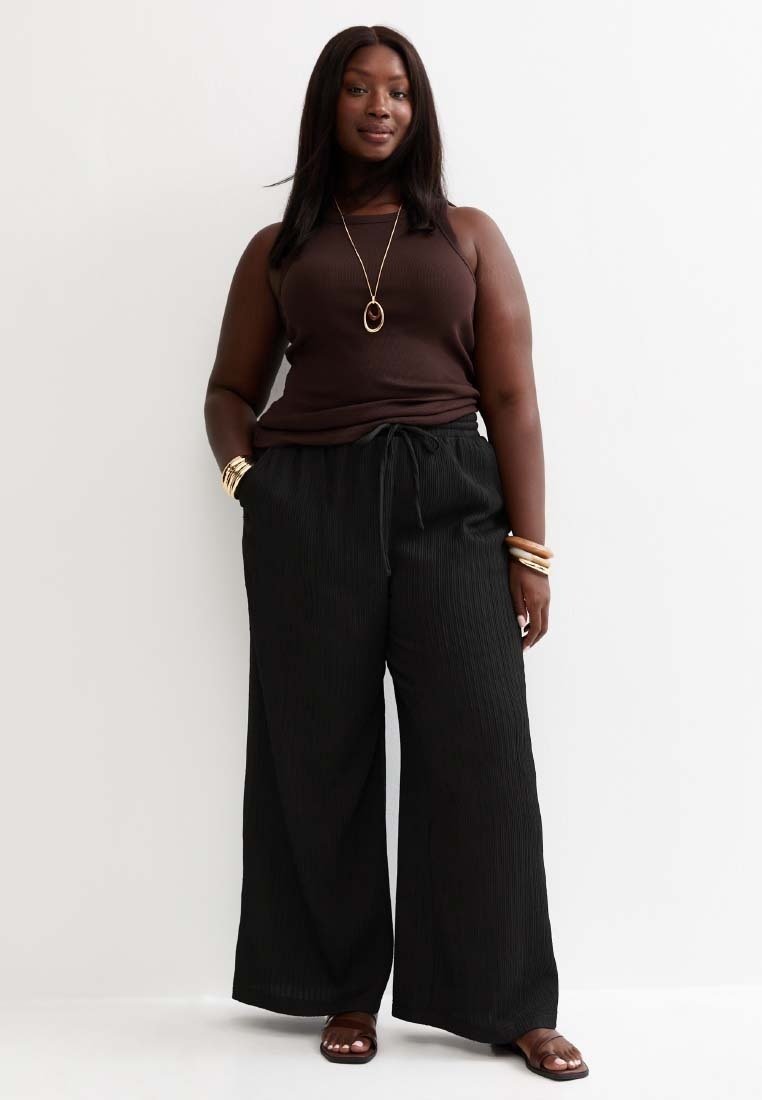 Mujer posa contra una pared blanca, vestida con un top sin mangas marrón oscuro, pantalones negros de pierna ancha, sandalias, collar y pulseras doradas.