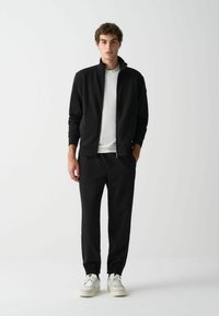 Fekete zipzáras dzseki magas gallérral, hozzáillő fekete melegítőnadrággal, rugalmas mandzsetta és fehér sportcipő. Egyszerű, minimalista dizájn.