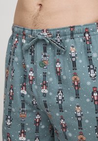 Men's pajama shorts in teal fabric featuring nutcracker-themed patterns, elastic waistband with drawstring, and a decorative green button.