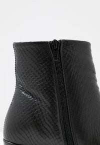 Detailní záběr na černou kotníkovou botu s texturovaným vzorem z umělé hadí kůže a viditelným černým zipem na boku.